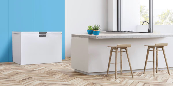 Μοντέρνα κουζίνα με ντουλάπια και νησίδα σε λευκό χρώμα, πλαισιωμένη από ξύλινο πάτωμα. Στη νησίδα υπάρχουν δύο ψηλά ξύλινα σκαμπό και δύο μικρές γλάστρες με παχύφυτα. Στο βάθος φαίνεται ένας ψυγείο-καταψύκτης μπροστά από έναν τοίχο με γαλάζια και λευκή απόχρωση. Η κουζίνα έχει μεγάλα παράθυρα με θέα προς έναν ηλιόλουστο εξωτερικό χώρο.